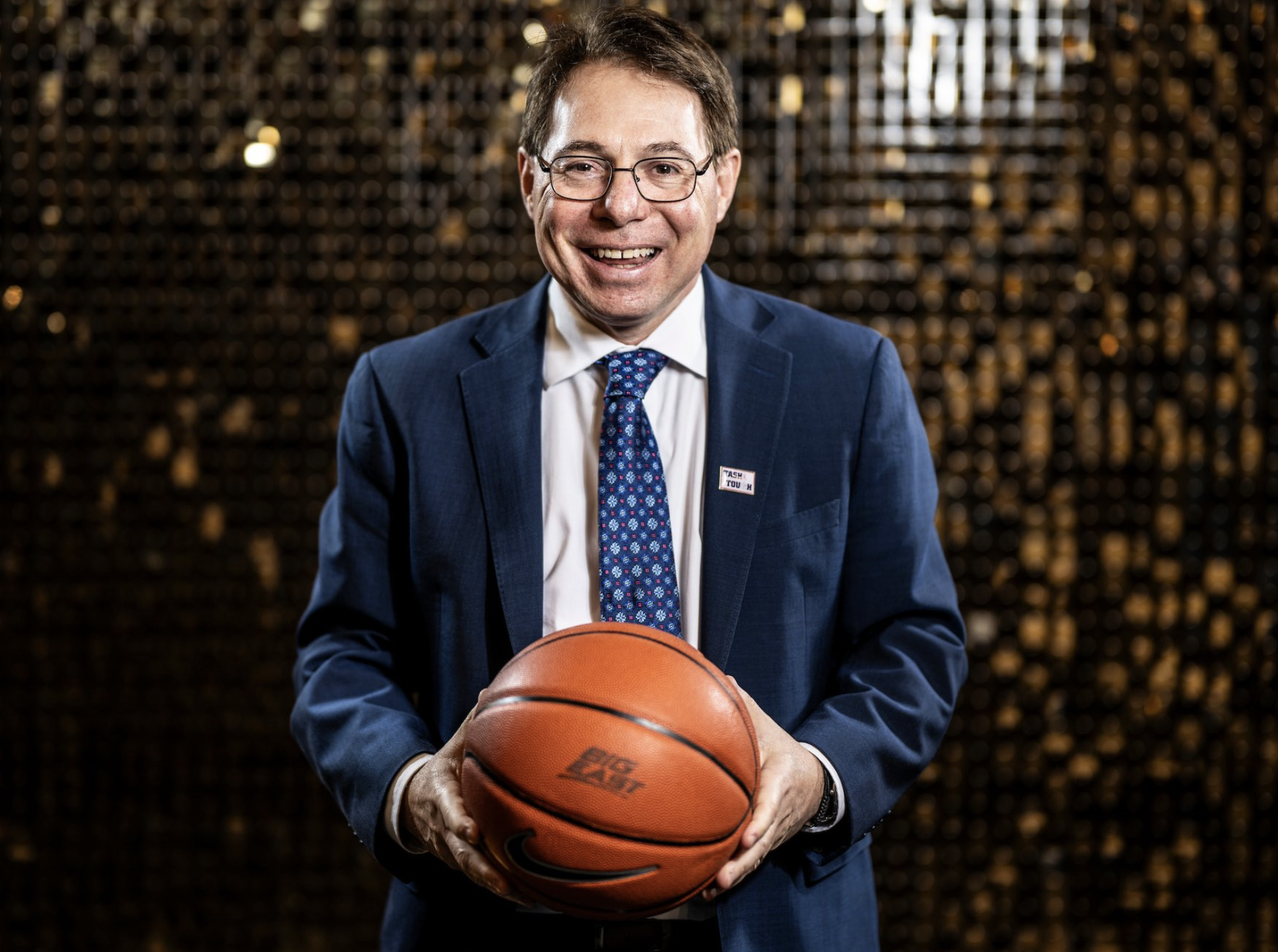 Seton Hall head coach Tony Bozzella is pictured in a suit and tie holding a BIG EAST-branded basketball. He is standing against a shimmering black and gold backdrop.
